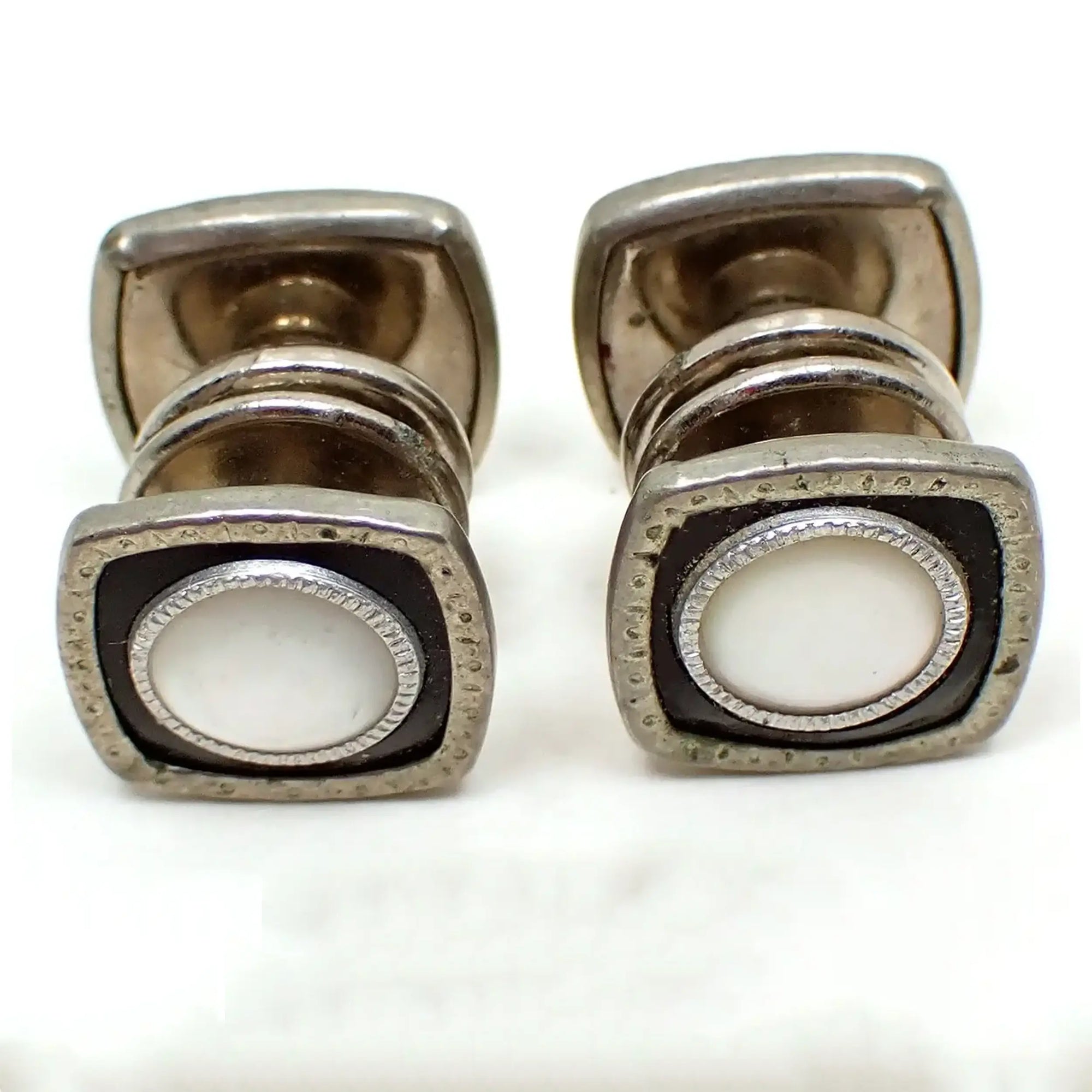 Enlarged angled view of the Art Deco snap together cufflinks. They are silver tone in color and are square in shape with rounded edges. There are black onyx gemstone fronts with a round area of mother of pearl shell in the middle. A few small dark spots can be seen on the outer edge of the metal and a small spot of verdigris can be seen where the two sides snap together in the middle.
