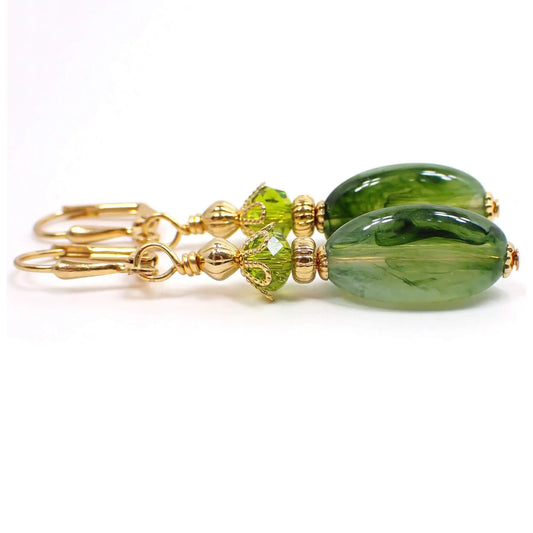 Side view of the handmade drop earrings. The metal is gold plated in color. There are new faceted glass crystal beads at the top in a peridot green color. The bottom beads are vintage lucite beads with marbled swirls of clear and shades of green. They are oval shaped with flat sides.