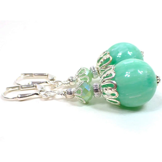Side view of the handmade drop earrings with vintage lucite beads. The metal is silver plated in color. There are pearly opaque faceted glass beads in a light green at the top. The bottom beads are round ball shaped lucite and are sea foam green with tiny lines of white color embedded in them.