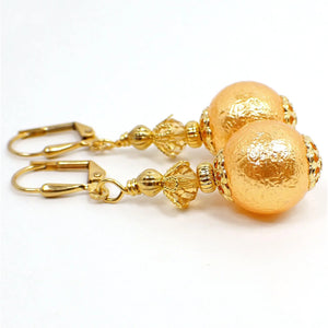 Side view of the handmade sphere drop earrings. The metal is gold plated in color. There is a light orange faceted glass crystal at the top. The bottom acrylic bead is round with a bumpy texture and is a peach light orange in color.