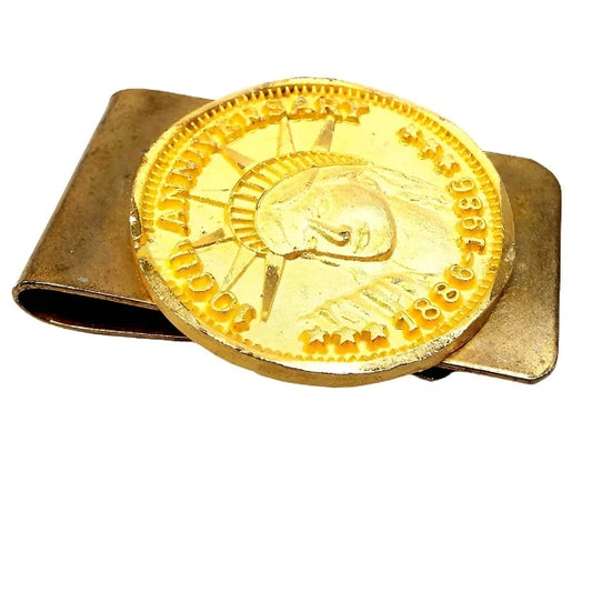 Angled top view of the retro vintage faux coin money clip. The clip base is a darkened gold tone in color from age. On the front is a bright gold tone color imitation coin that has the Statue of Liberty head and the words 100th Anniversary 1886-1986. The edges of the faux coin are faceted.