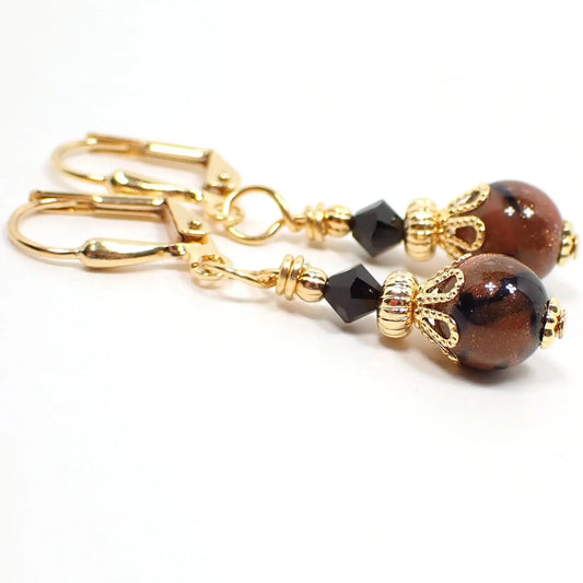 Photo of the handmade goldstone drop earrings. The metal is gold plated in color. There are faceted black crystal glass bicone beads at the top and small round ball goldstone beads at the bottom. The goldstone beads are marbled with orange glass that has tiny flecks of copper and black.
