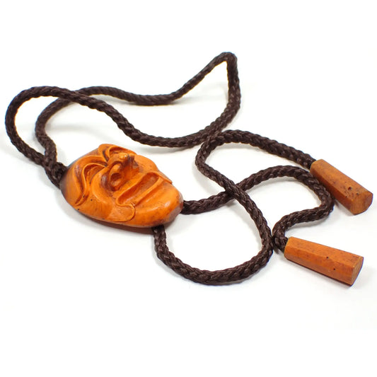 Top view of the retro vintage South Korean Hahoetal theater mask bolo tie. The cord is dark brown and thick braided. There is a carved smiling theater mask as the bolo slide. The ends are carved resin as well and cone shaped. The slide and ends are mostly orange in color with some areas of dark brown.