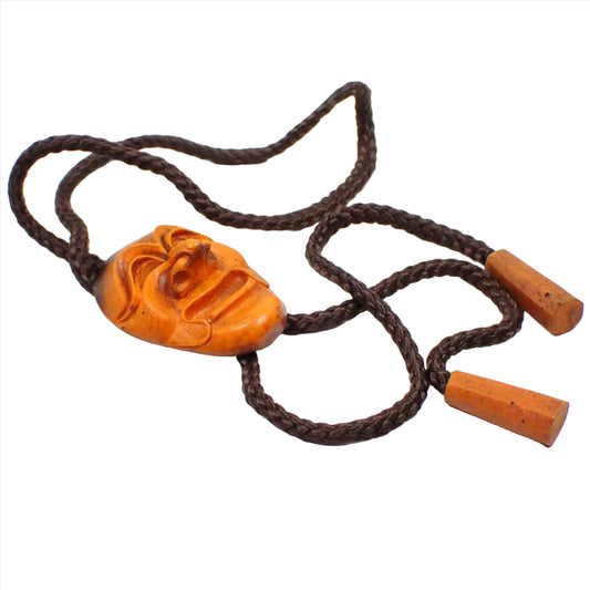 Top view of the retro vintage South Korean Hahoetal theater mask bolo tie. The cord is dark brown and thick braided. There is a carved smiling theater mask as the bolo slide. The ends are carved resin as well and cone shaped. The slide and ends are mostly orange in color with some areas of dark brown.