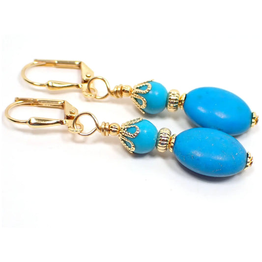 Photo of the handmade magnesite and chalk turquoise drop earrings. The metal is gold plated in color. There are small round blue dyed magnesite beads at the top. The bottom beads are puffy oval shaped and are chalk turquoise which is ground down, dyed, and then added to resin to form the beads.
