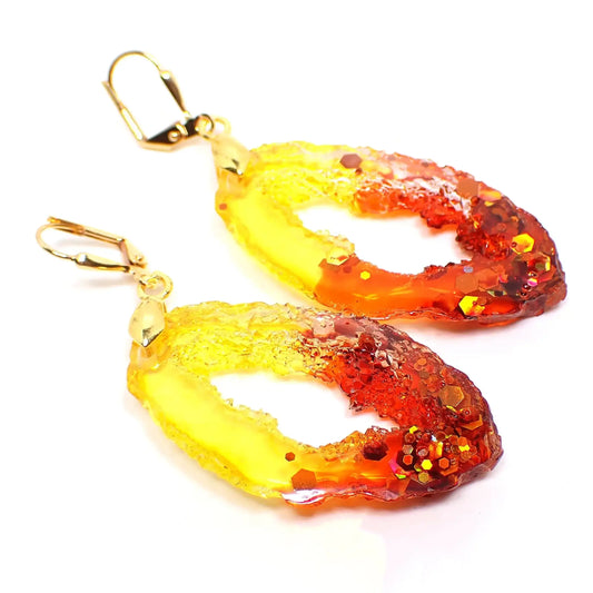 Angled front view of the handmade faux druzy geode slice style earrings. The metal is gold plated in color. The resin is fiery red and orange towards the bottom and bright yellow at the top. There is chunky iridescent glitter at the bottom with shades of orange and yellow.