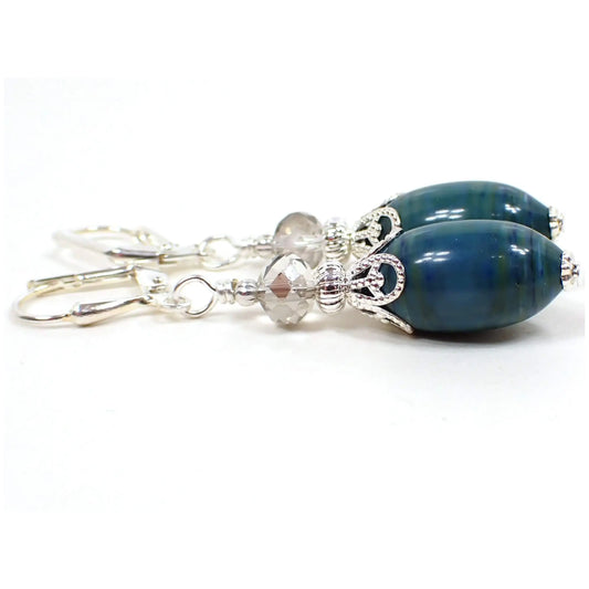 Side view of the handmade earrings with vintage lucite beads. The metal is silver plated in color. There are faceted glass crystal beads at the top in a very light gray color. The bottom lucite beads are oval shaped and have marbled areas with different shades of blue and green. Each beads has a different pattern for a unique appearance.