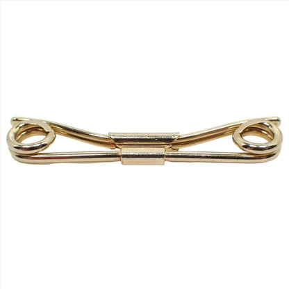Enlarged front view of the Mid Century vintage Swank collar clip. It is gold tone plated in color. The front has a rounded bar that has spiral ends on each side. The back has a curved design with two thinner rounded bars together.