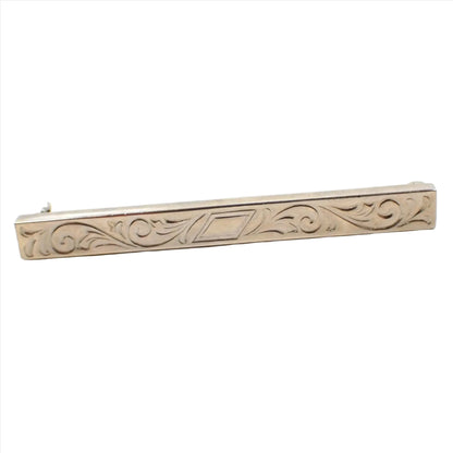 Enlarged front view of the Mid Century vintage bar brooch. It has a longer rectangle shape with an angled rectangle in the middle and scroll like leaf designs on either side. The metal is a slightly darkened silver tone color.