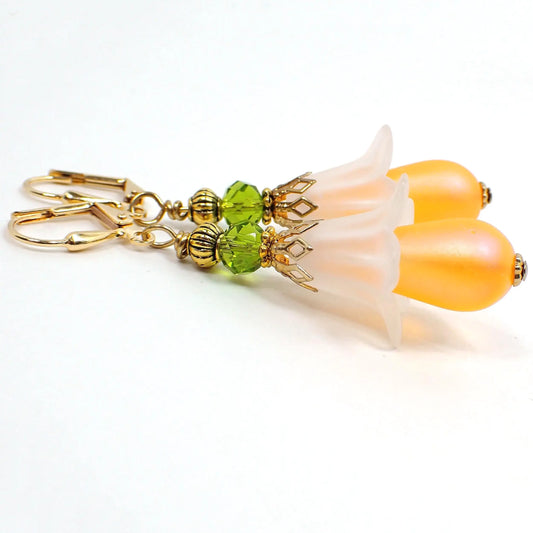 Angled view of the handmade flower earrings. The metal is gold plated in color. There is a faceted green glass crystal bead at the top. The flower petal bead is made of lucite and is a frosted matte white color. The bottom bead is a vintage German acrylic teardrop bead in matte frosted orange.