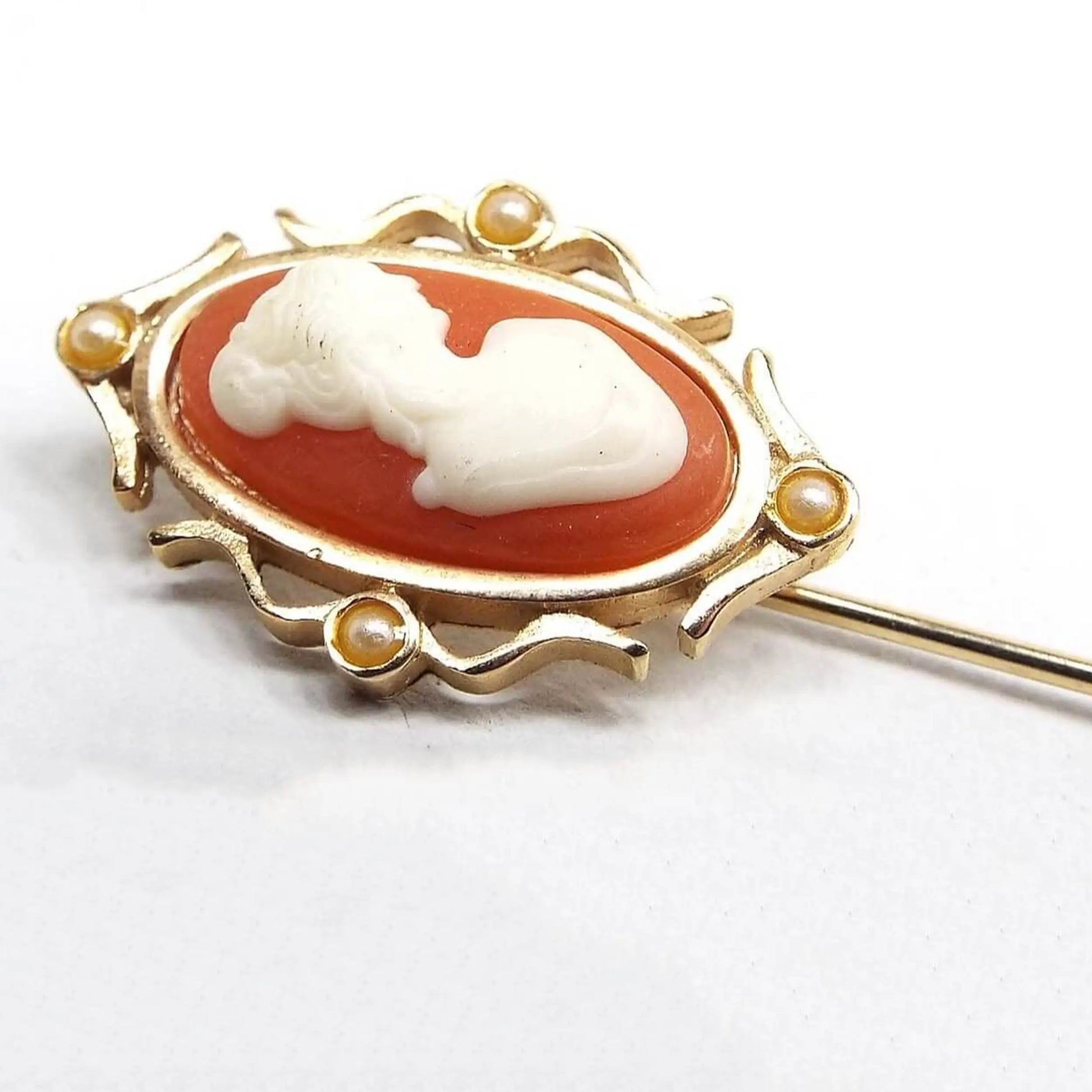 Magnified view of the top part of the Avon retro vintage cameo stick pin. The metal is gold tone in color. It has plastic cameo bust of a woman on a salmon orange background. The top is oval with a metal fancy curved frame on each side with an off white faux pearl on each side.
