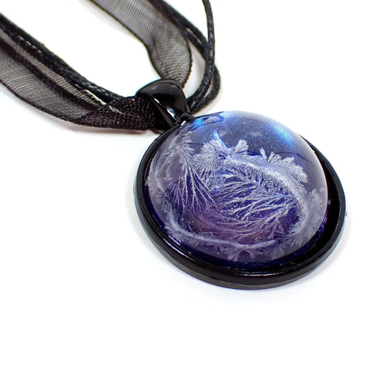 Enlarged view of the handmade black and resin pendant necklace. The necklace has three black strands of faux leather cord and a strand of organza ribbon. The pendant has a black coated base and is round. There is a domed round cab on the front made of resin with an iridescent blue background and a crystalline frost like design going through it.