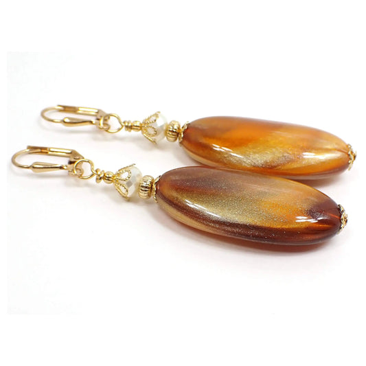 Angled view of the big handmade marbled lucite oval drop earrings. The metal is gold plated in color. There are shimmery off white faceted glass beads at the top. The bottom vintage lucite beads have marbled shades of caramel brown and metallic gold.