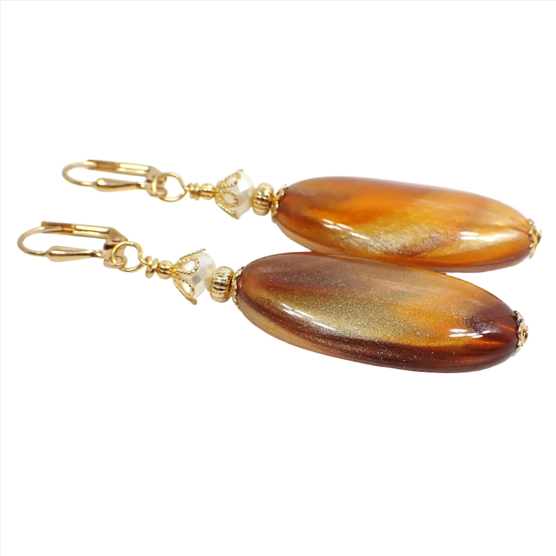 Angled view of the big handmade marbled lucite oval drop earrings. The metal is gold plated in color. There are shimmery off white faceted glass beads at the top. The bottom vintage lucite beads have marbled shades of caramel brown and metallic gold.