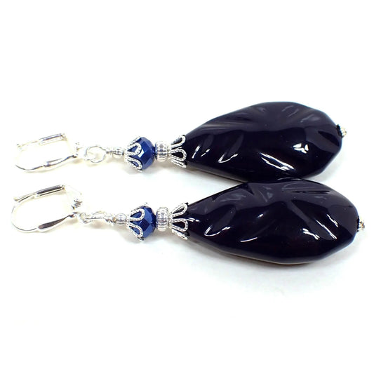 Angled view of the large handmade teardrop earrings with vintage lucite beads. The metal is silver plated in color. There are metallic blue faceted glass beads at the top. The bottom vintage lucite beads are large puffy flat teardrop shaped with an indented pattern and are dark blue in color.