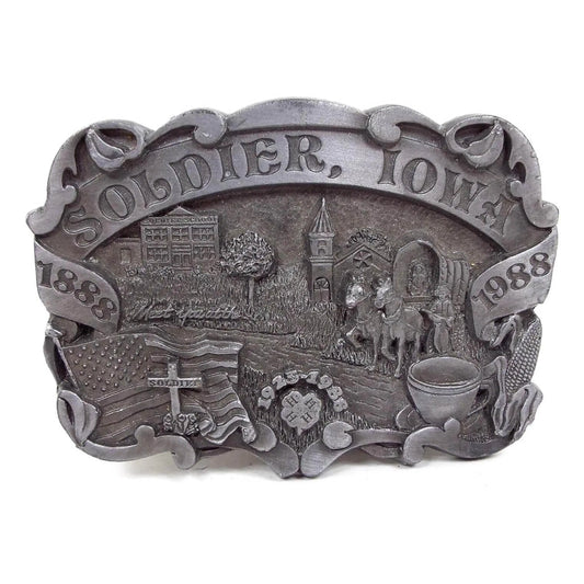 Front view of the Dart Inc 4H Belt Buckle. It is gray in color. The top says Soldier, Iowa. It has a school building, church, US Flag, 4H symbol, cup and saucer, corn, covered wagon with horses, and a cross gravestone marker on the front.