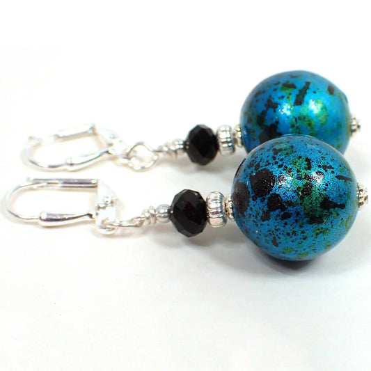 Side view of the handmade drop earrings. The metal is silver plated in color. There are black faceted glass beads at the top. The bottom beads are acrylic and are round ball shaped in a bright metallic teal blue color with black and green splash pattern. Each bottom bead is different from the other in pattern for a unique look.