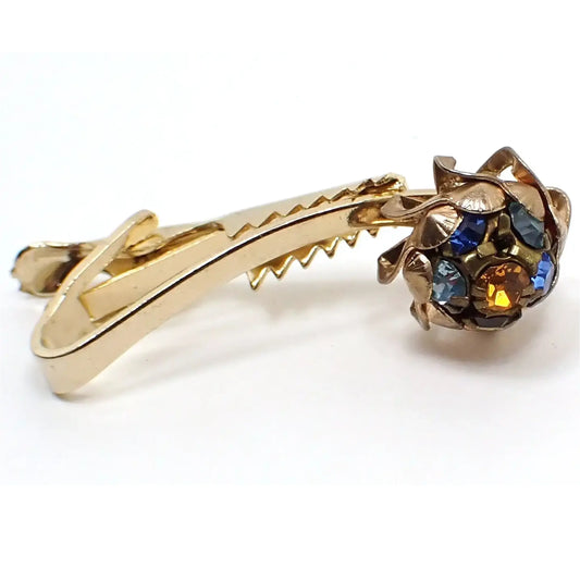 Angled front view of the 1950s Mid Century vintage rhinestone tie clip. The metal is gold tone plated in color.  The bar in the front is arced and has a pinwheel style shape at the end with a cluster of different colored rhinestones. There are four shades of blue rhinestones, a purple rhinestone, and an orange rhinestone. 