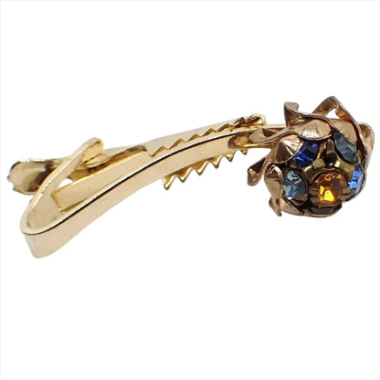Angled front view of the 1950s Mid Century vintage rhinestone tie clip. The metal is gold tone plated in color.  The bar in the front is arced and has a pinwheel style shape at the end with a cluster of different colored rhinestones. There are four shades of blue rhinestones, a purple rhinestone, and an orange rhinestone. 