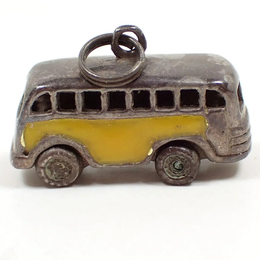Enlarged side view of the retro vintage bus charm.  It's shaped like a school bus or transportation bus. The sterling silver is darkened from age. There is yellow enamel on the sides. The wheels are on small metal axels and can rotate. There is a split jump ring at the top of the charm.