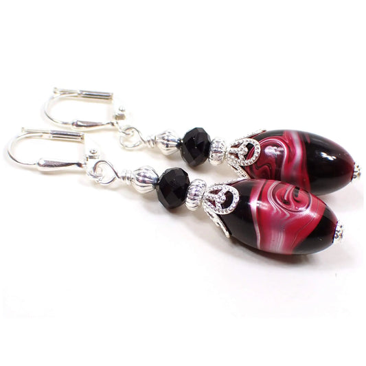 Angled view of the handmade earrings with vintage lucite beads. The metal is silver plated in color. There are black faceted glass beads at the top. The bottom lucite beads have marbled swirls of bright pink, metallic silver, and black. Each bottom bead is different in pattern from the other for a unique appearance.