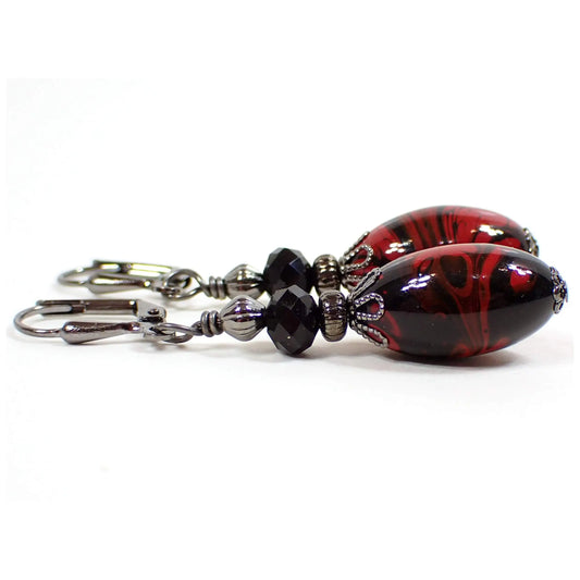 Side view of the handmade Goth drop earrings with vintage lucite beads. The metal is gunmetal gray in color. There is a faceted glass black bead at the top. The bottom oval beads are vintage lucite and have a swirled red and black pattern on them. The marbled pattern on each bottom bead is different from the other.