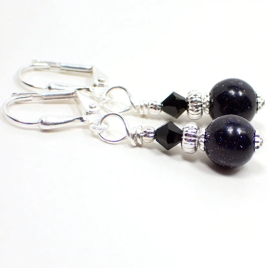 Angled view of the small handmade blue goldstone drop earrings. The metal is silver plated in color. There is a small faceted black crystal glass bead at the top. The bottom bead is round ball shaped and is blue goldstone, which is a dark blue glass with fine and tiny blue and silver flecks of metallic glitter.