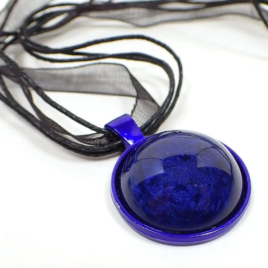 Enlarged close up of the handmade goth resin necklace. The necklace part has black organza with is like a semi transparent ribbon. There are also three strands of black waxed cord. The pendant is round and bright blue enameled metal. The front of the pendant has a round domed handmade cab that has pearly blue resin that goes from dark blue to a vivid, almost cobalt blue in color.