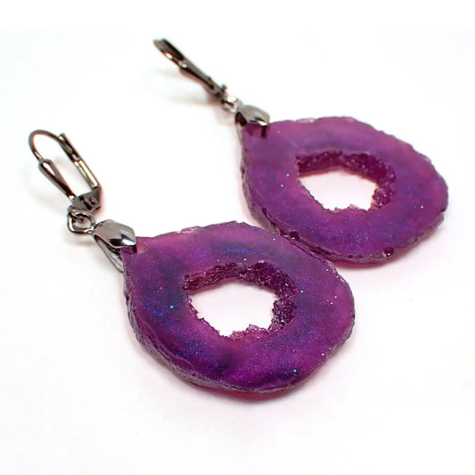 Angled front view of the handmade geode slice style earrings. The metal is dark gunmetal gray in color. They are shaped like rounded teardrops with a freeform edge. The resin is a dark pearly purple color with hints of blue in the light.