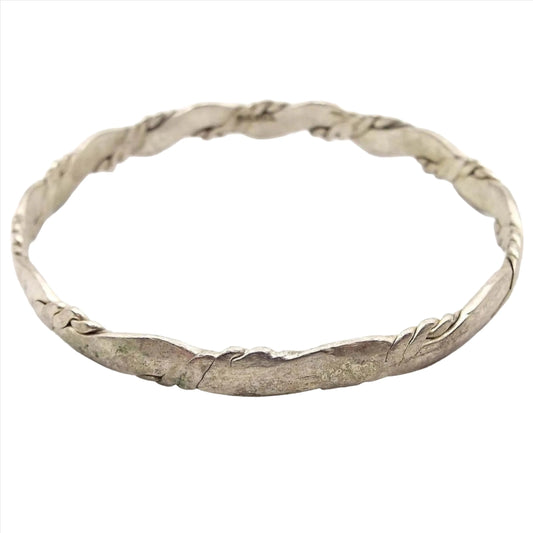 Angled view of the retro vintage Taxco bangle bracelet. It has a twisted design that's been hammered so the inside and outside area are flat. It is a darkened silver tone in color.