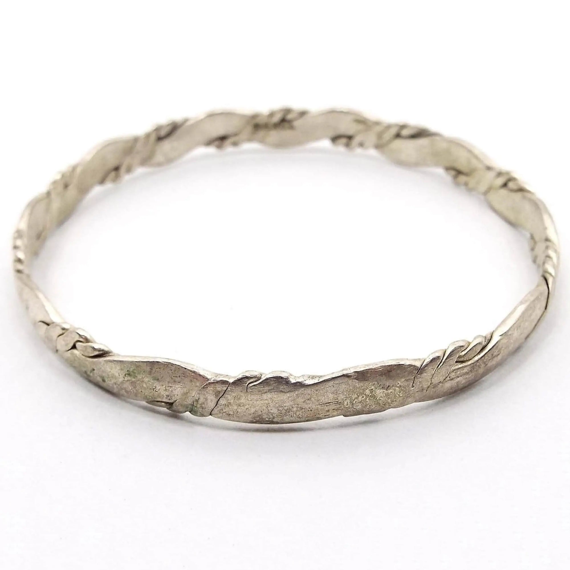 Angled view of the retro vintage Taxco bangle bracelet. It has a twisted design that's been hammered so the inside and outside area are flat. It is a darkened silver tone in color.