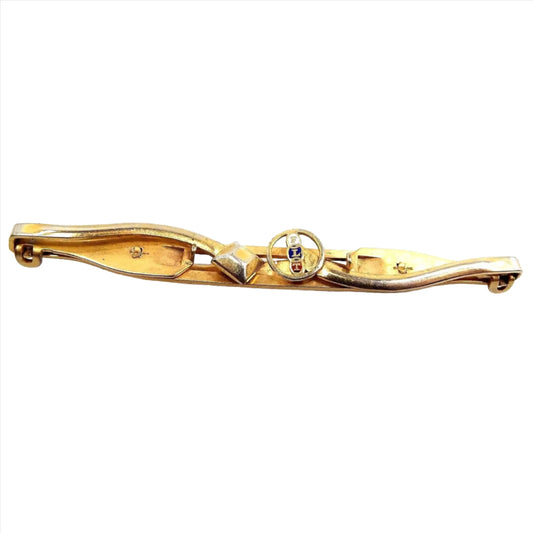 Top view of the Mid Century vintage Anson FLT Odd Fellows tie bar. It's long gold tone in color with each side that opens out as a clip. The left side has a diamond shape at the end and the right side has an open circle with FLT going down the middle of it. The letters are enameled with white, dark blue, and red. 