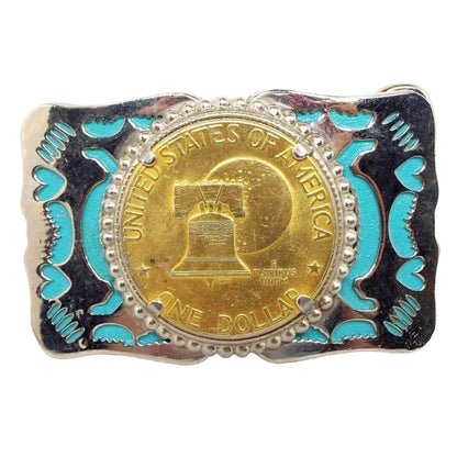 Front view of the retro vintage belt buckle. It is silver tone in color that has an abstract and heart cut out areas with blue enamel inside. The middle has a gold tone color replica dollar coin of the Liberty Bell and moon.