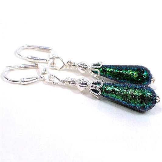 Angled side view of the handmade glitter earrings with vintage German acrylic beads. The metal is silver plated in color. The bottom acrylic beads are teardrop shaped and are black with green glitter on the outside.