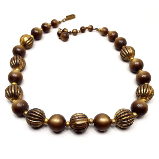 Top view of the Mid Century vintage moonglow lucite and antiqued brass necklace. The beads are round in shape. The brass beads are dark brass in color and are corrugated with lines. The moonglow lucite beads are a golden brown in color and the hues shift with a glowy appearance as you move around. There is a hook clasp at the end.