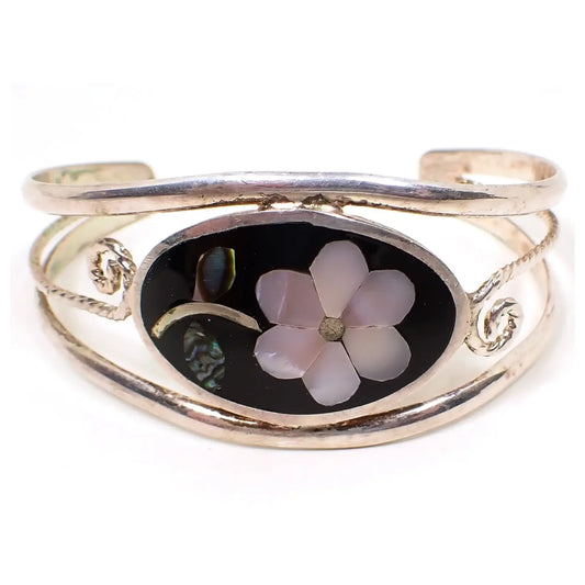 Front view of the retro vintage Mexican floral cuff bracelet, The metal is silver tone in color. There is an oval on the front with black enamel and a flower design that has inlaid pieces of mother of pearl shell for the petals and abalone shell for the leaves.