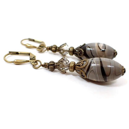 Side view of the handmade gray oval lucite drop earrings. The bottom beads are oval lucite plastic and are marbled with swirled shades of gray, brown, black, and thin bands of white. The bead on each earring looks different from the other. The top beads are faceted glass crystal in a very light gray, almost clear color. The metal is antiqued brass in color.