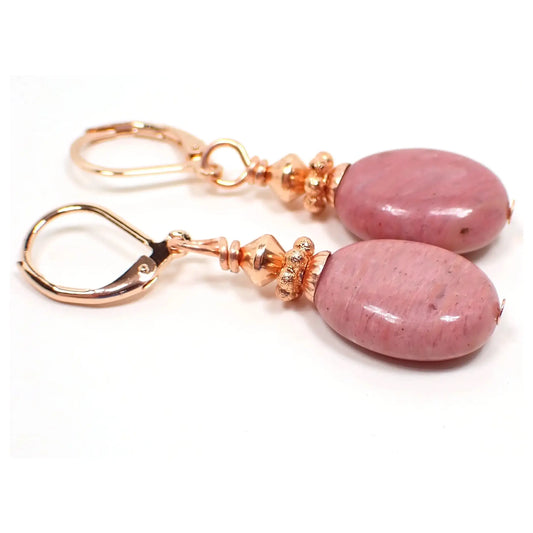 Angled view of the handmade rhodonite earrings. The photo shows rose gold plated lever backs at the top and copper plated beads at the top of the drops. The gemstones are puffy oval shaped and have shades of soft rosy pink.