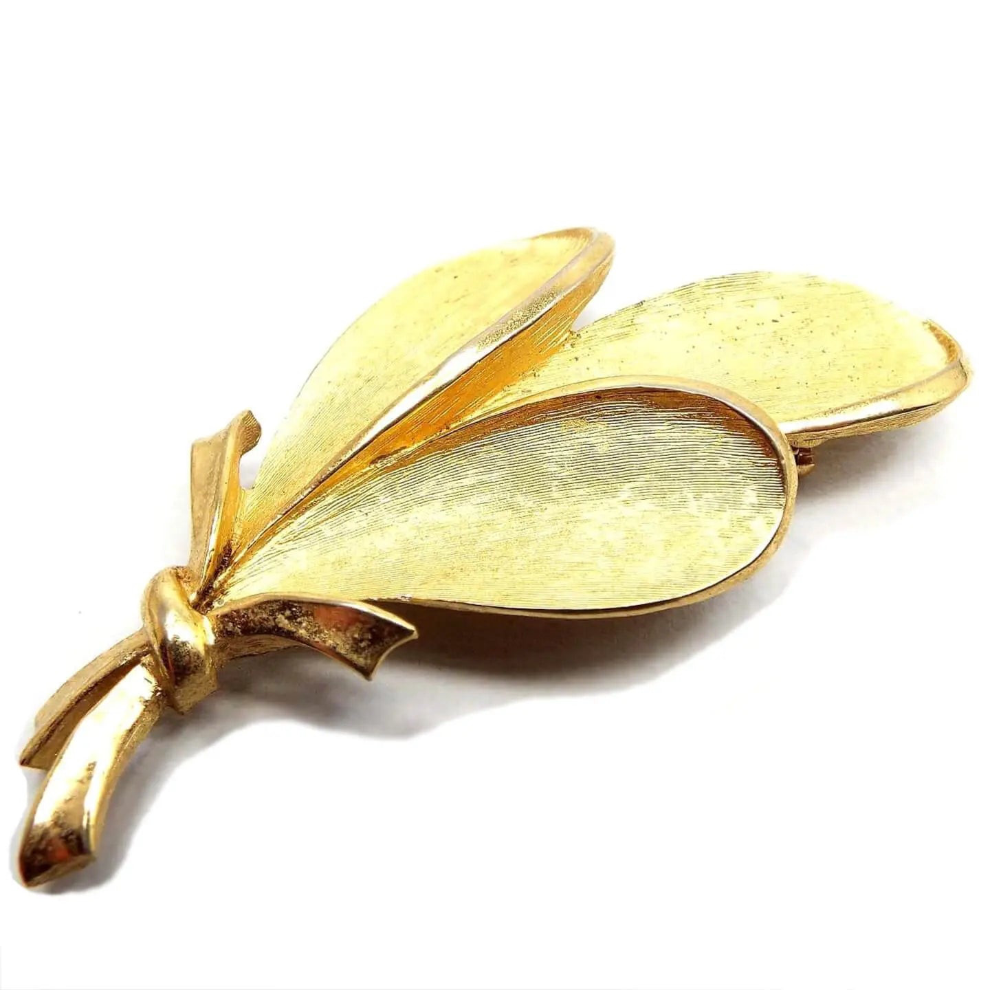 Front view of the Mid Century vintage Crown Trifari brooch pin. The metal is gold tone in color and has a matte brushed appearance. There are three leaves with teardrop shaped ends that look like they're tied together at the stems with a ribbon.
