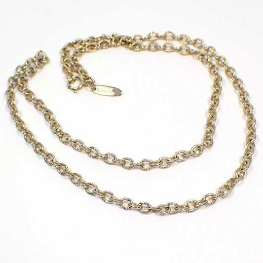 Angled view of the retro vintage Whiting and Davis chain necklace. The metal is gold tone in color. There are smaller sized oval cable chain links. The links have a textured pattern stamped on them. There is a spring ring clasp at the end and an oval hang tag with Whiting Davis on it.