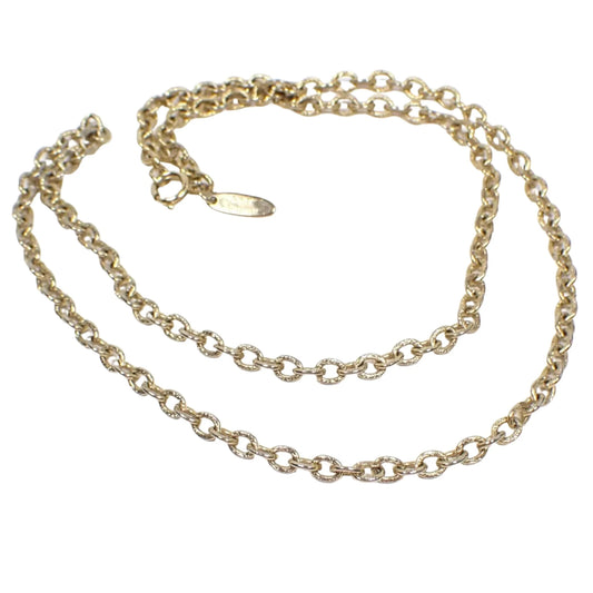 Angled view of the retro vintage Whiting and Davis chain necklace. The metal is gold tone in color. There are smaller sized oval cable chain links. The links have a textured pattern stamped on them. There is a spring ring clasp at the end and an oval hang tag with Whiting Davis on it.