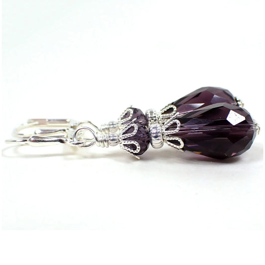 Side view of the handmade drop earrings. The metal is silver plated in color. There are dark purple faceted glass crystal rondelle beads on the top and teardrop shaped beads on the bottom.