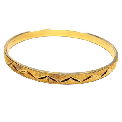 Angled side view of the Mid Century vintage Crown Trifari bangle. The bracelet is gold tone in color and has a thin line and angled V cut design all the way around the edge.
