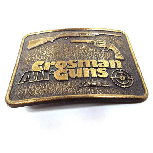 Angled front view of the retro vintage Crosman Air Guns Belt Buckle.. It is antiqued brass in color and has a rifle style and handheld style air gun at the top. There is a bullseye in the bottom right corner where it's marked Coleman company.