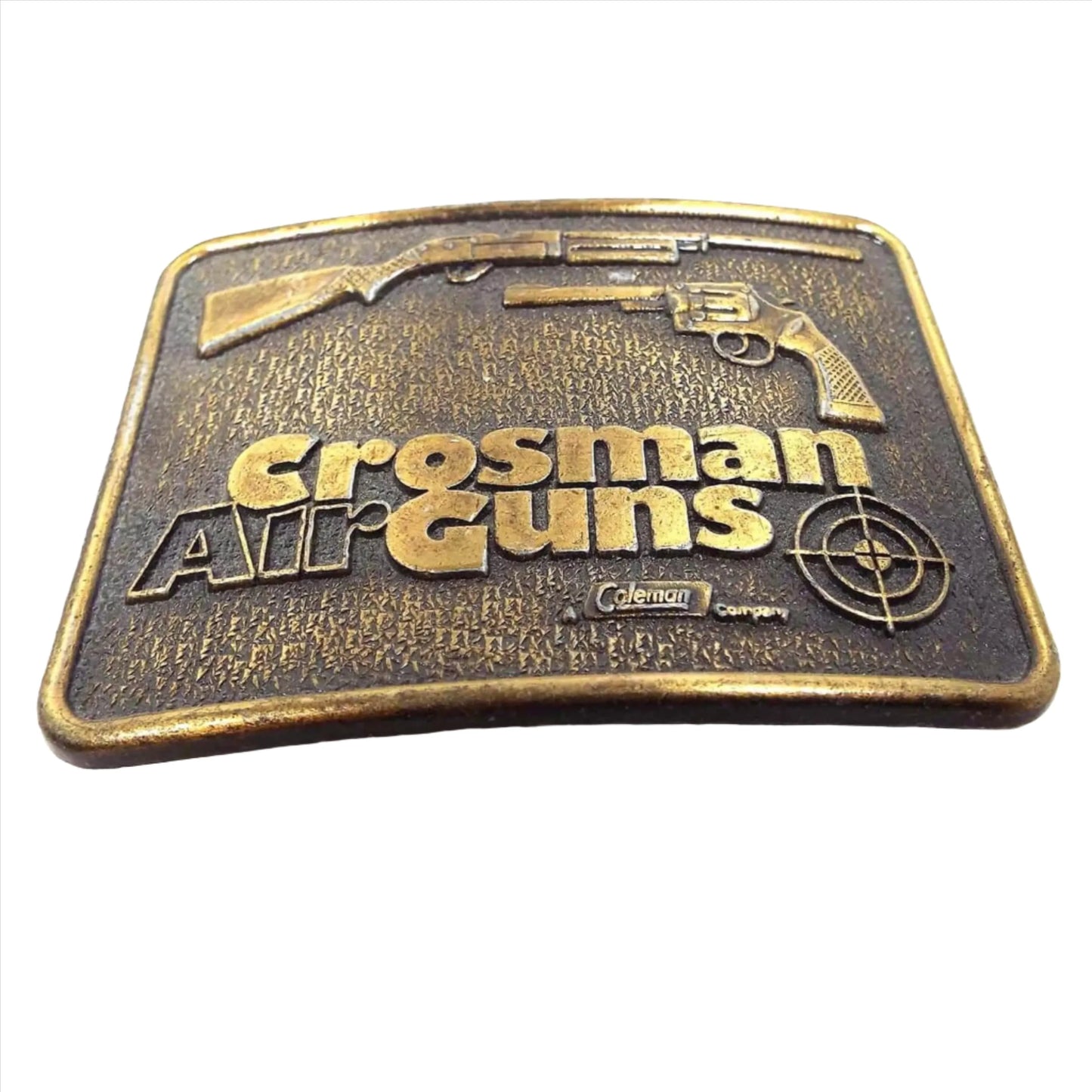 Angled front view of the retro vintage Crosman Air Guns Belt Buckle.. It is antiqued brass in color and has a rifle style and handheld style air gun at the top. There is a bullseye in the bottom right corner where it's marked Coleman company.