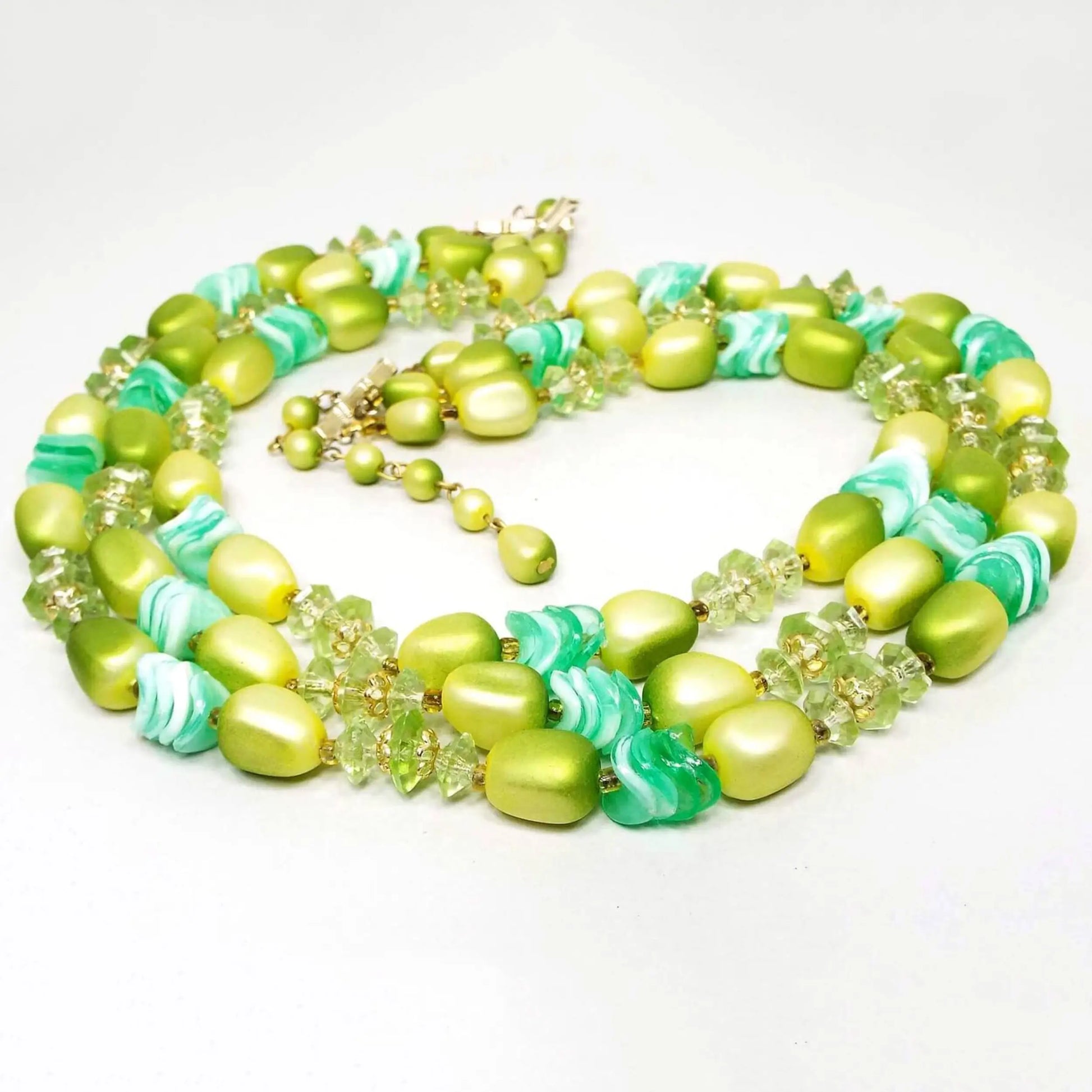The Mid Century vintage beaded multi strand necklace from Hong Kong. It has three strands of plastic beads in varying shades of bright to medium green. It has a fairly chunky appearance with disc, saucer, nugget, and round shaped beads.