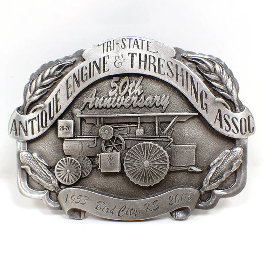 Front view of the pre owned modern Antique Engine and Threshing Association belt buckle. It's made of pewter and a gray in color. The association's name is at the top and then it shows 50th anniversary and 1953 Bird City, KS 2003. There is an old piece of farm equipment depicted in the middle with corn and wheat at the corners.