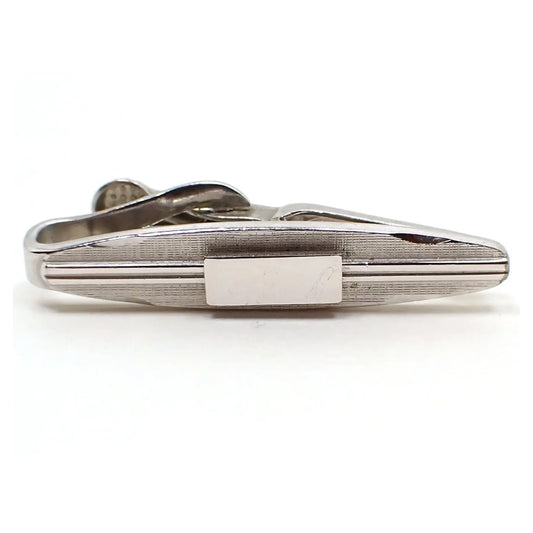 Front view of the small Swank Mid Century vintage tie clip. It is a small oval shape with silver tone color metal. The front has a beveled edge on each side and a textured matte design. In the middle are two lines on either side going towards a middle rectangle area. The stripes and rectangle are shiny silver tone in color.