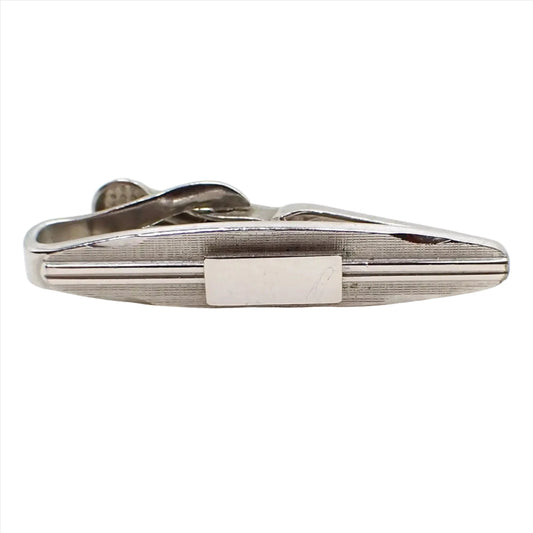 Front view of the small Swank Mid Century vintage tie clip. It is a small oval shape with silver tone color metal. The front has a beveled edge on each side and a textured matte design. In the middle are two lines on either side going towards a middle rectangle area. The stripes and rectangle are shiny silver tone in color.