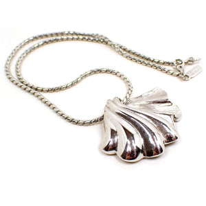 Front view of the retro vintage Monet pendant necklace. The metal is silver tone in color. There is a snake chain with a hinged clasp and Monet hang tag at the end. The pendant is a fan seashell shape with some shiny silver and some textured silver areas.